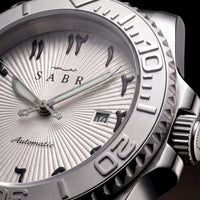 Sabr automatic watch with a silver sunburst dial, black Arabic numeral hour markers, a textured stainless steel bezel, and a matching metal bracelet, shown against a dark background.