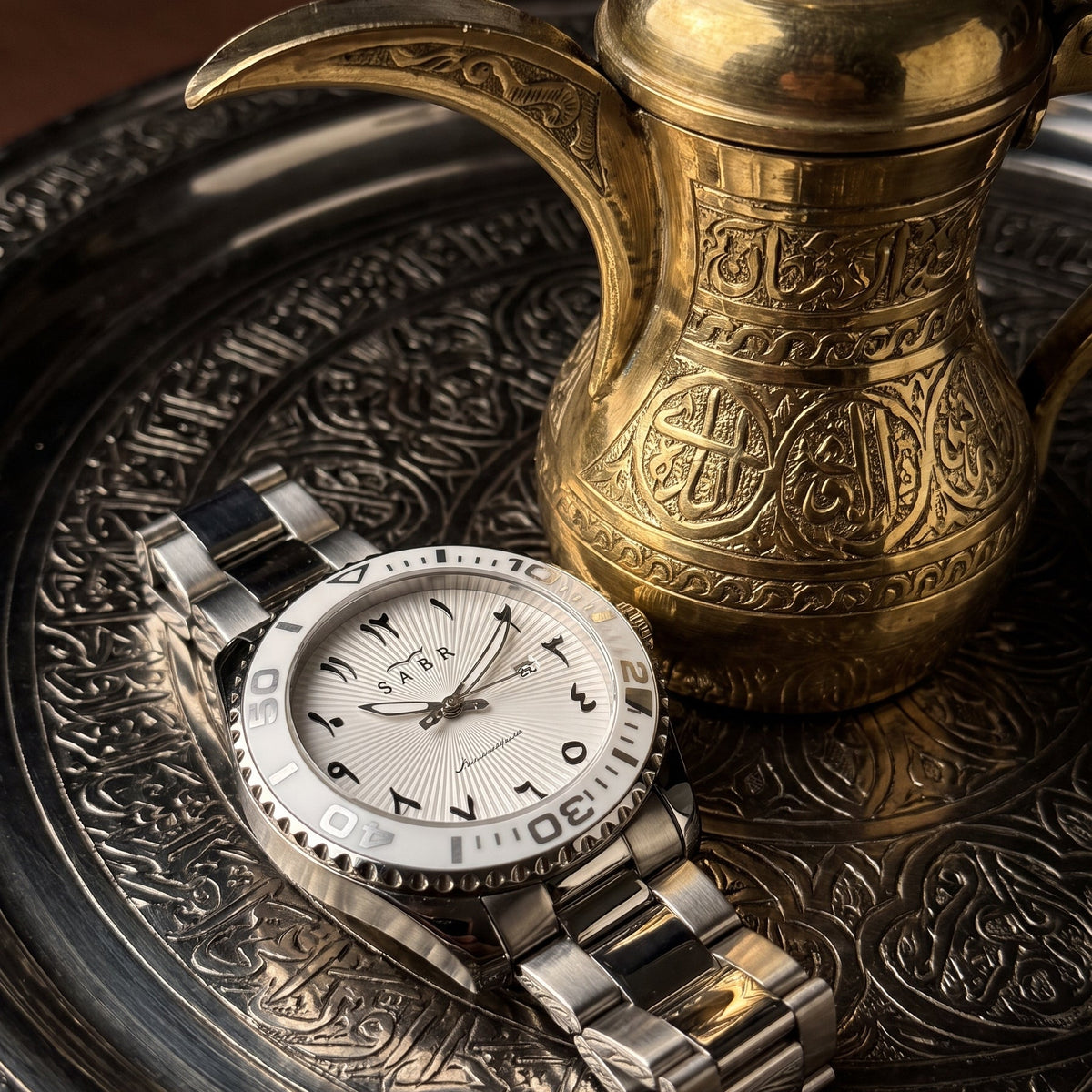 A stainless steel and white dial wristwatch with a bezel, styled on an ornate silver tray next to a gold Arabic coffee pot.