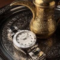 A stainless steel and white dial wristwatch with a bezel, styled on an ornate silver tray next to a gold Arabic coffee pot.